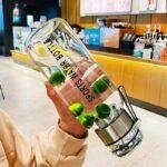 Main tenant une gourde transparente avec infuseur et morceaux de citron vert dans un café