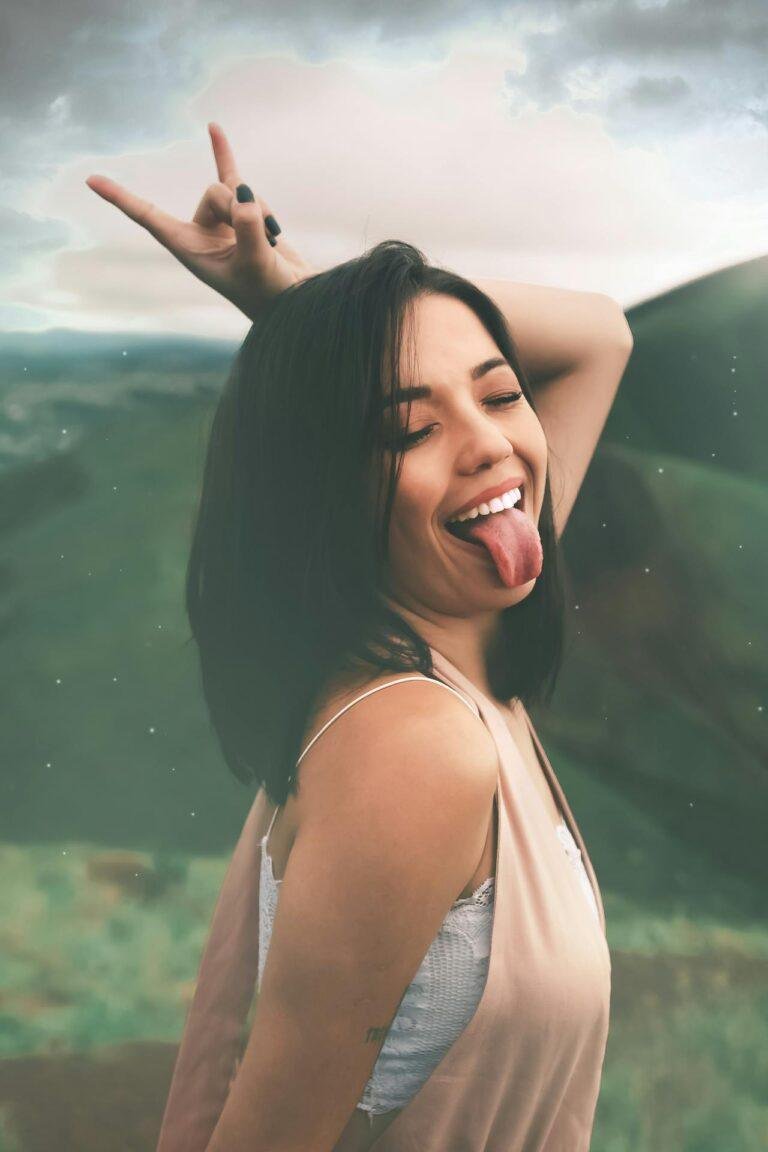 Jeune femme souriante tirant la langue avec légèreté dans un paysage naturel, symbole de plaisir et de complicité lors d’un cunnilingus respectueux et attentif.