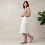 Femme de profil portant des bottes santiags roses à talon bas, une jupe midi satin blanche et un pull beige, dans un intérieur minimaliste avec plante verte.